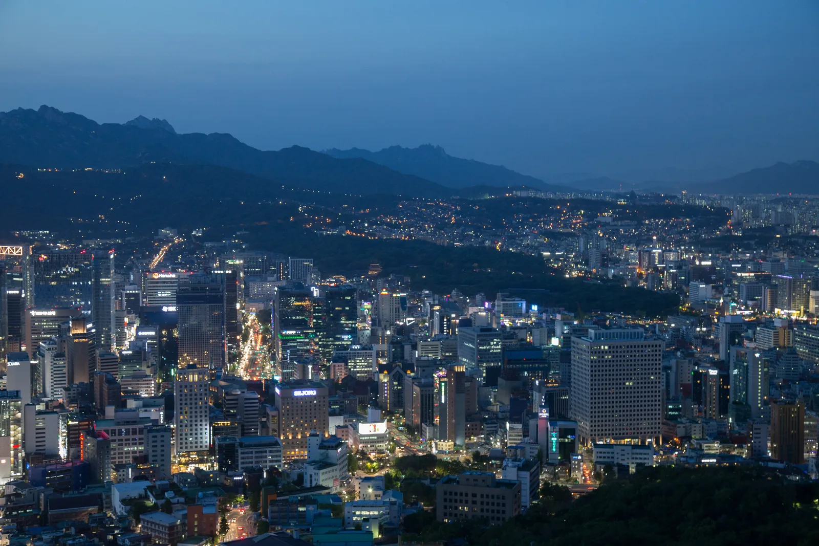 A wide evening view of central Seoul with city lights and mountains in the distance.